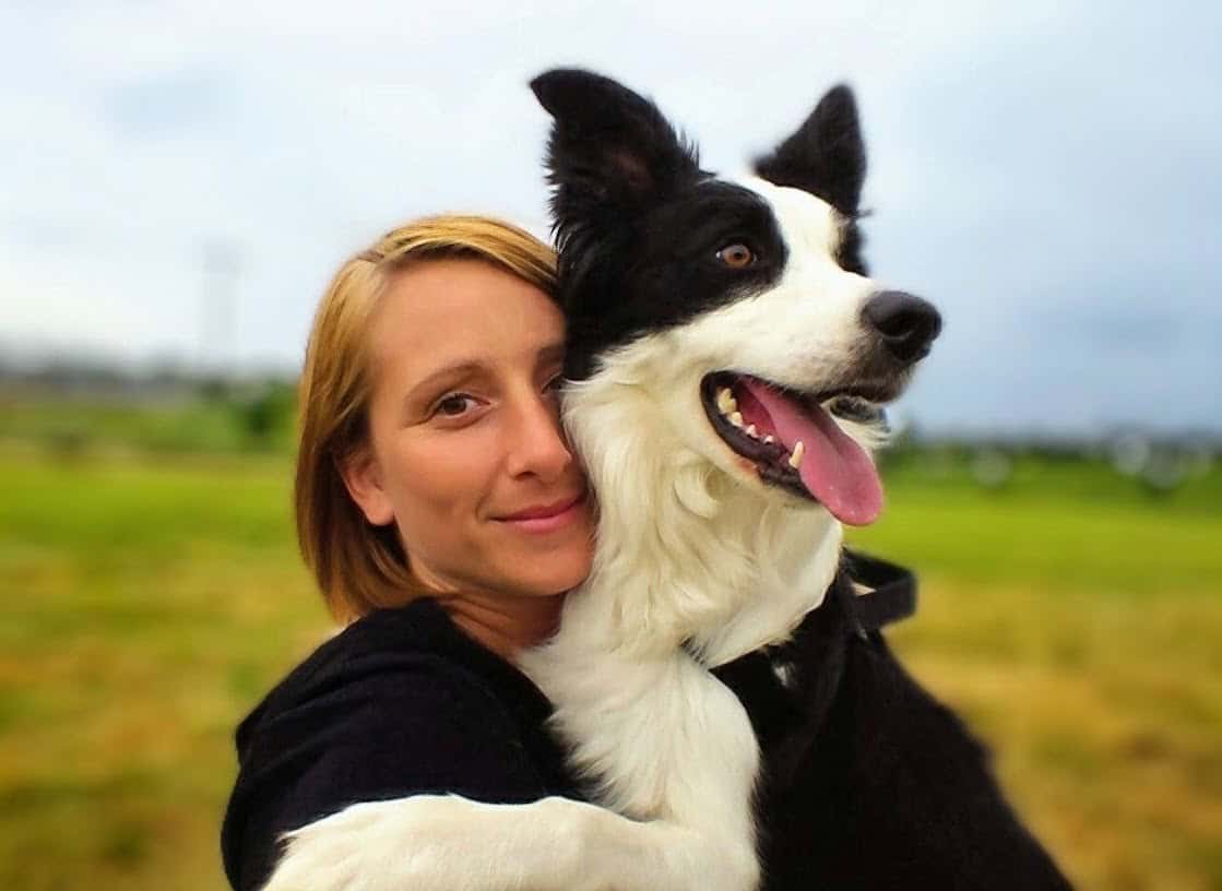 Frau umarmt ihren schwarz-weißen Hund auf einer Wiese – beide wirken glücklich und verbunden.